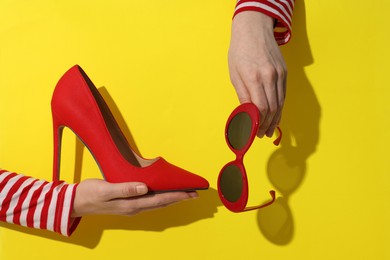 Photo of Woman showing red high-heeled shoe and sunglasses on yellow background, closeup