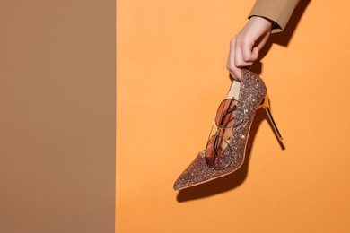 Photo of Woman showing glitter high-heeled shoe and sunglasses on color background, closeup. Stylish presentation