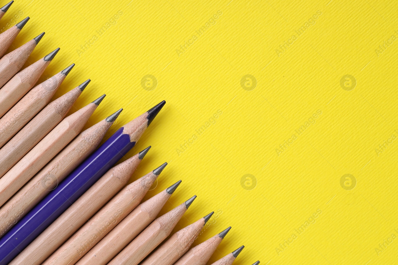 Photo of One blue pencil among graphite ones on yellow background, top view. Space for text