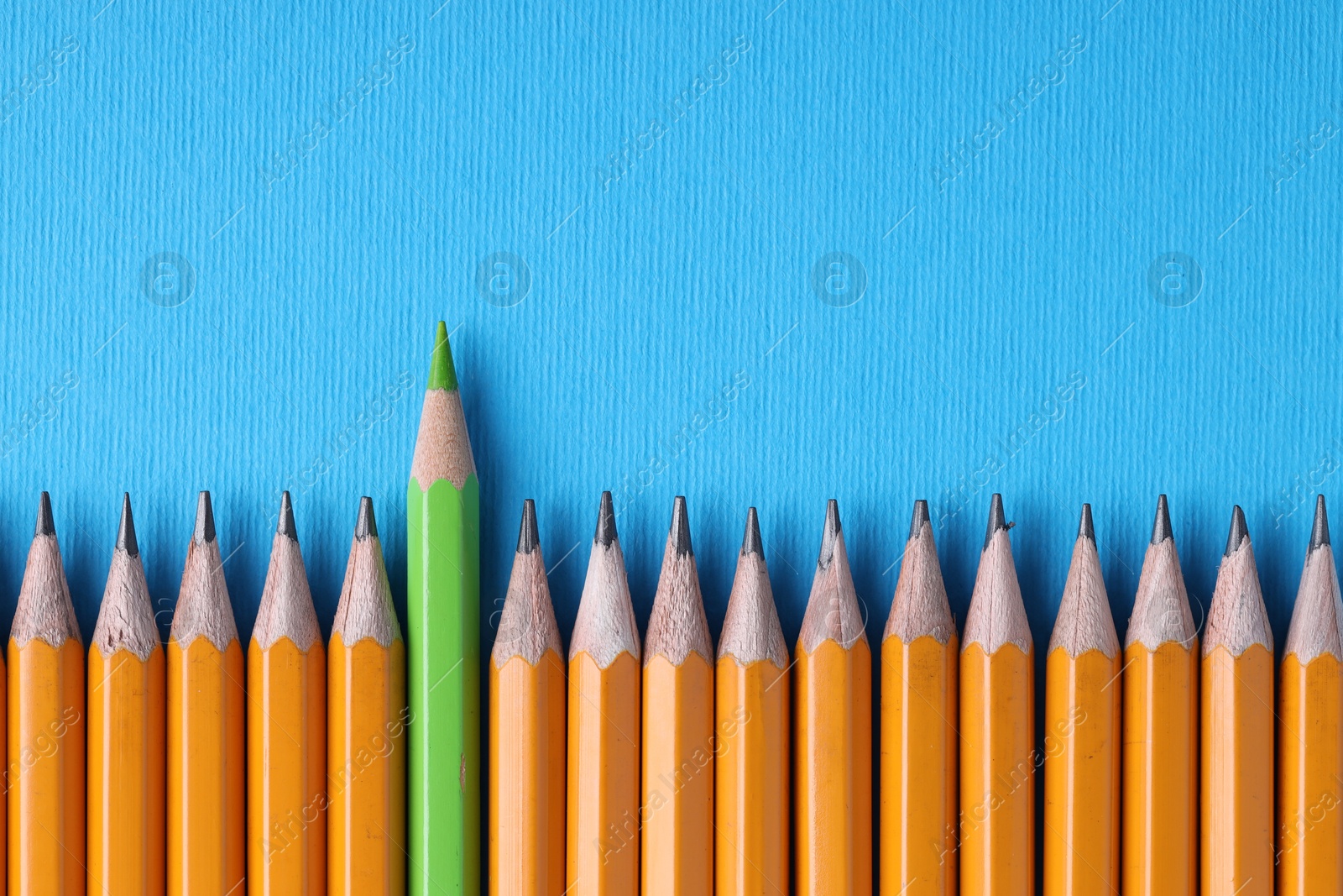 Photo of One green pencil among graphite ones on light blue background, top view. Space for text