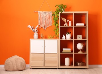 Photo of Wooden shelving unit with different home decor, houseplant and pouf near orange wall indoors