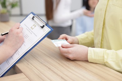 Patient with medical insurance card and receptionist filling document at wooden counter in clinic, closeup