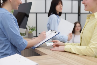 Patient with medical insurance card and receptionist filling document at wooden counter in clinic, closeup