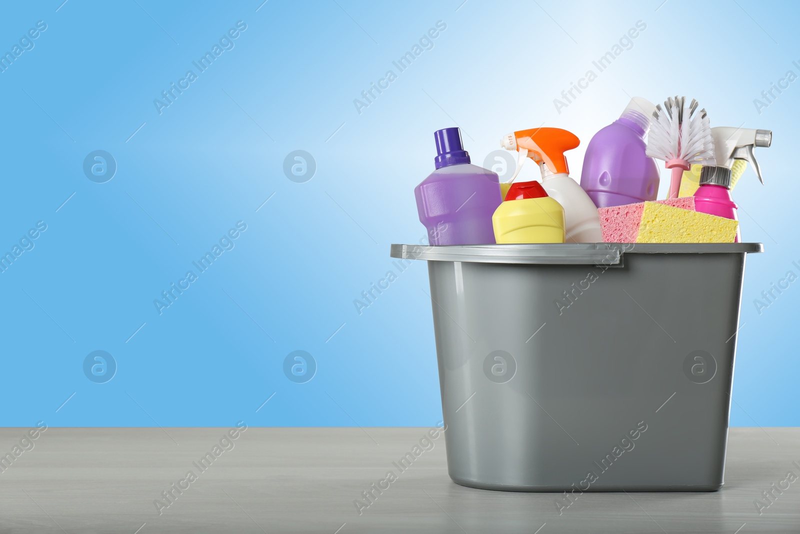 Image of Different cleaning products and supplies in bucket on wooden table against light blue gradient background, space for text