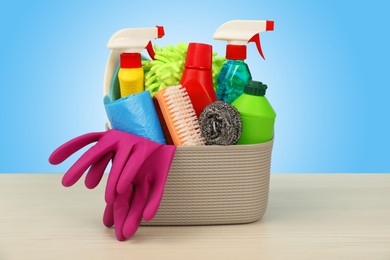 Different cleaning products and supplies in basket on wooden table against light blue gradient background