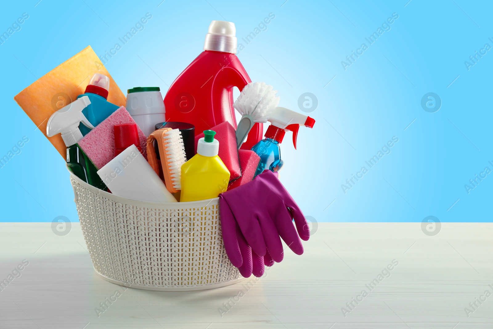 Image of Different cleaning products and supplies in basket on wooden table against light blue gradient background, space for text