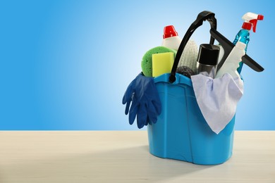 Image of Different cleaning products and supplies in bucket on wooden table against light blue gradient background, space for text