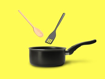 Image of Slotted spatula and wooden spoon in air over saucepan on yellow background