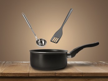 Image of Slotted spatula and ladle in air over saucepan on wooden table against pale brown background