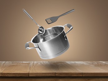 Saucepan, ladle and slotted spatula in air over wooden table against pale brown background