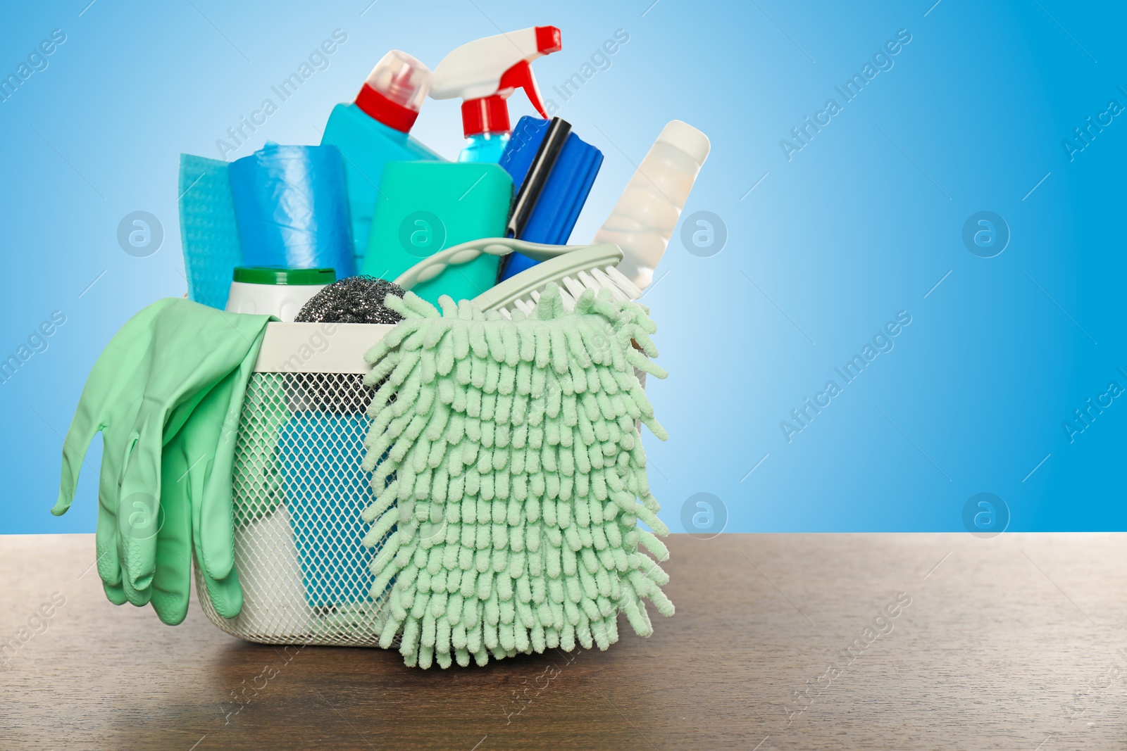 Image of Different cleaning products and supplies in basket on wooden table against light blue gradient background, space for text