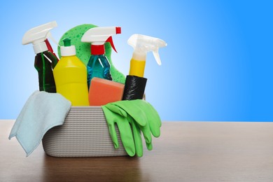 Image of Different cleaning products and supplies in basket on wooden table against light blue gradient background, space for text