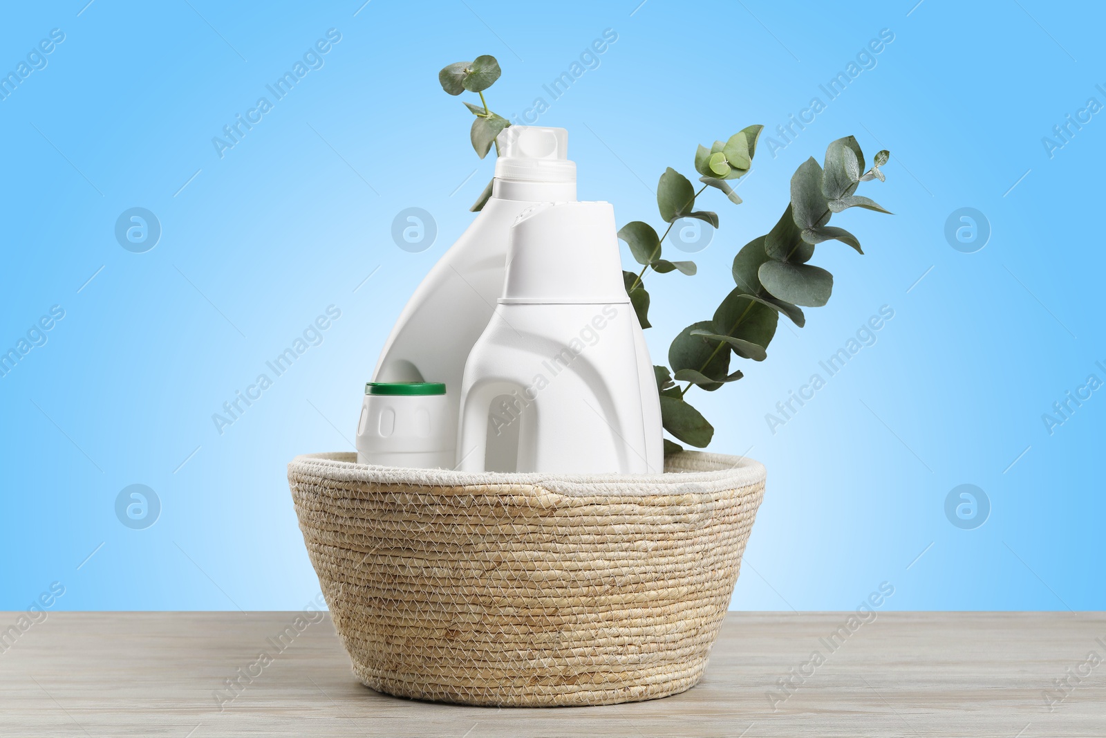 Image of Eco-friendly cleaning products and eucalyptus branches in wicker basket on wooden table against light blue gradient background