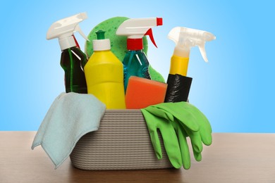 Image of Different cleaning products and supplies in basket on wooden table against light blue gradient background