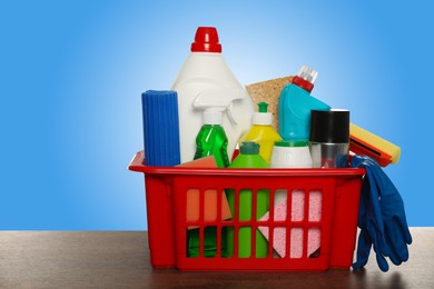 Image of Different cleaning products and supplies in basket on wooden table against light blue gradient background, space for text