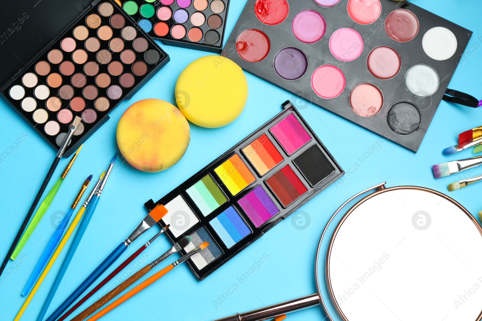 Photo of Bright face paints, brushes, mirror and sponges on light blue background, flat lay