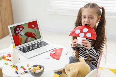 Photo of Little girl with paper fly agaric at white table indoors. Child creativity and craft