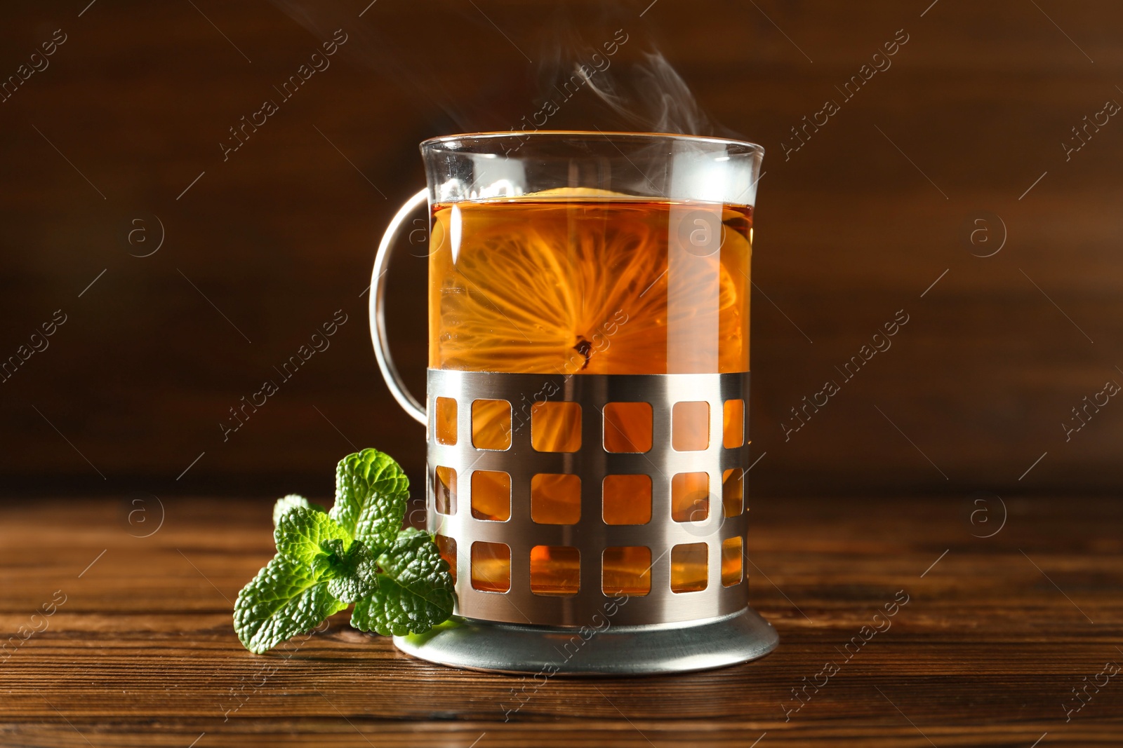 Photo of Glass of hot tea with lemon in holder and mint on wooden table, closeup