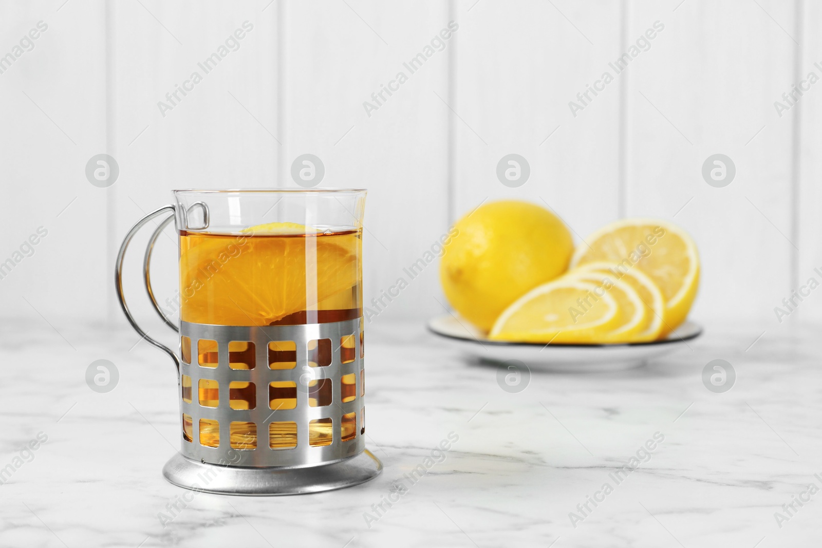 Photo of Glass cup of tea in metal holder and lemon slices on white marble table, closeup