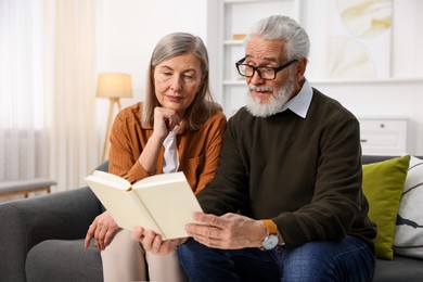 Photo of Cute elderly couple reading book together on sofa at home