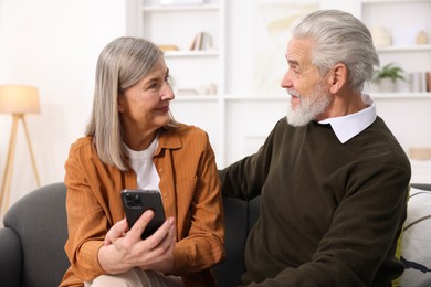 Cute elderly couple with smartphone looking at each other on sofa at home