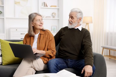 Photo of Happy elderly couple with laptop looking at each other on sofa at home