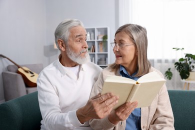 Photo of Cute elderly couple reading book together on sofa at home