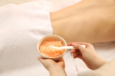 Photo of Esthetician applying cosmetic product for body wraps treatment onto woman's back in spa salon, closeup