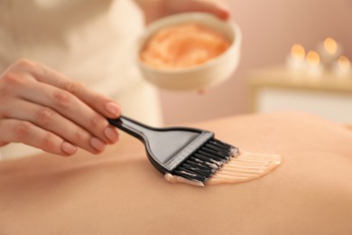 Esthetician applying cosmetic product for body wraps treatment onto woman's back in spa salon, closeup