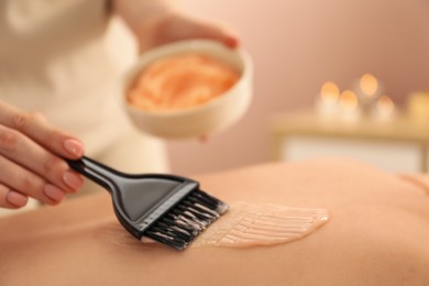 Esthetician applying cosmetic product for body wraps treatment onto woman's back in spa salon, closeup