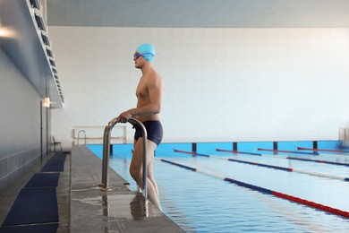 Photo of Young man in cap, goggles and swimwear getting out of swimming pool indoors, space for text