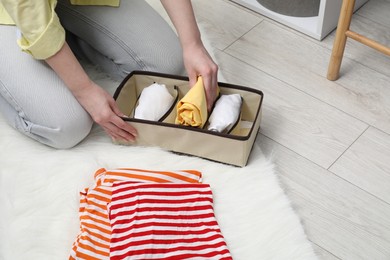 Photo of Woman putting folded clothes into organizer on floor at home, closeup