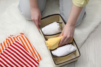 Photo of Woman putting folded clothes into organizer on floor at home, above view