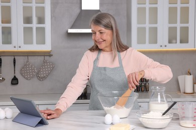 Photo of Senior woman reading recipe on tablet while cooking in kitchen. Online culinary book