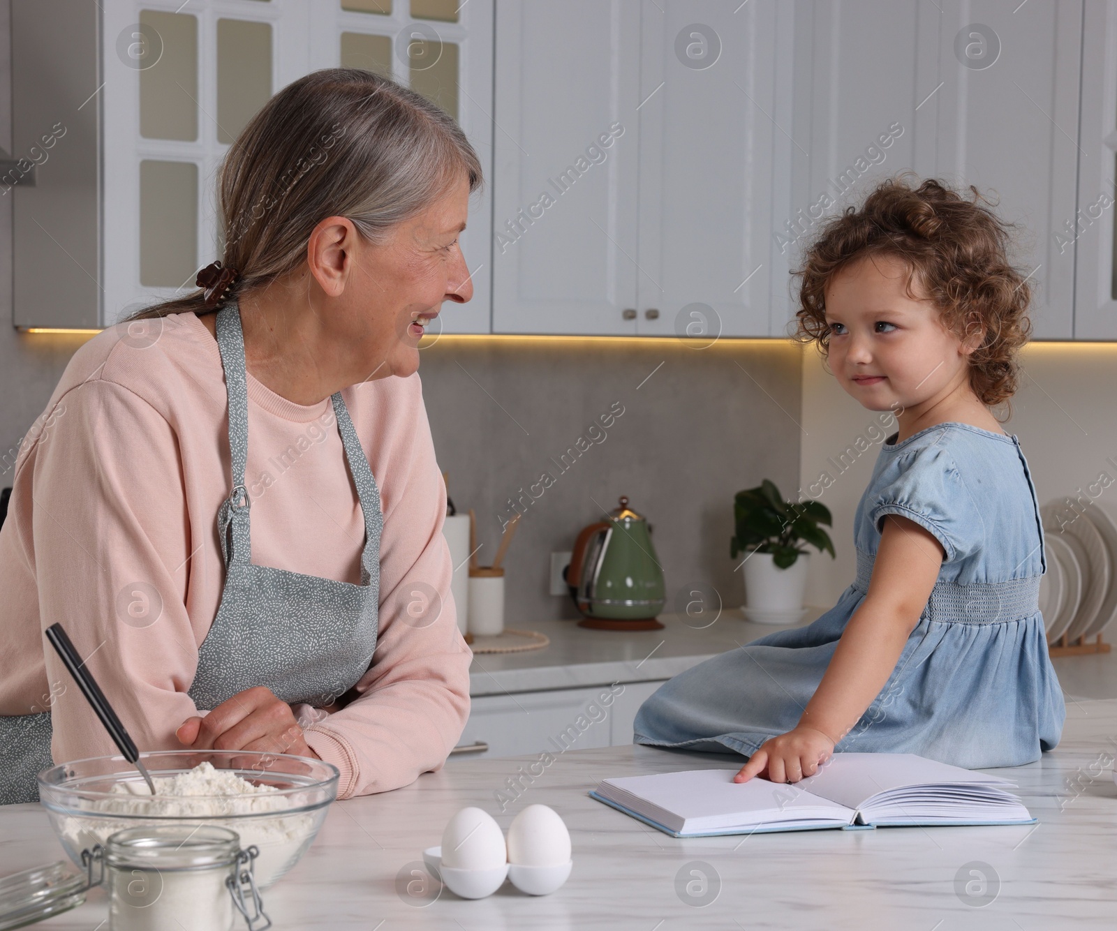 Photo of Cute little girl with her granny cooking by recipe book in kitchen