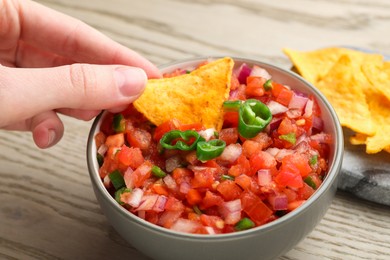 Photo of Woman dipping nacho chip into delicious salsa sauce at wooden table, closeup