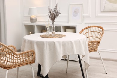 Photo of Table with white tablecloth, dried flowers in vases and chairs in stylish room. Interior design