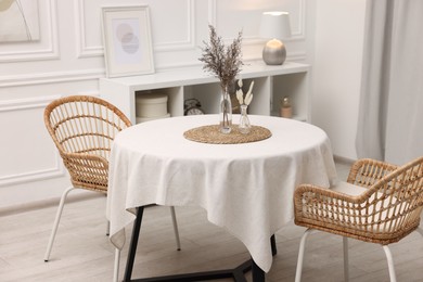 Photo of Table with white tablecloth, dried flowers in vases and chairs in stylish room. Interior design