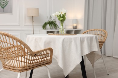 Photo of Table with white tablecloth, flowers in vase and chairs in stylish room. Interior design