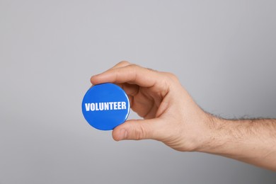 Photo of Man holding button badge with word Volunteer on light grey background, closeup