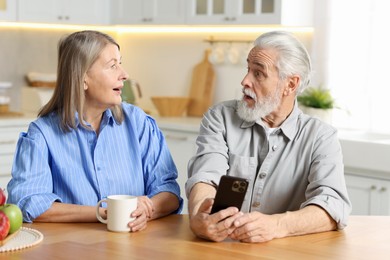 Surprised elderly couple with smartphone at table in kitchen