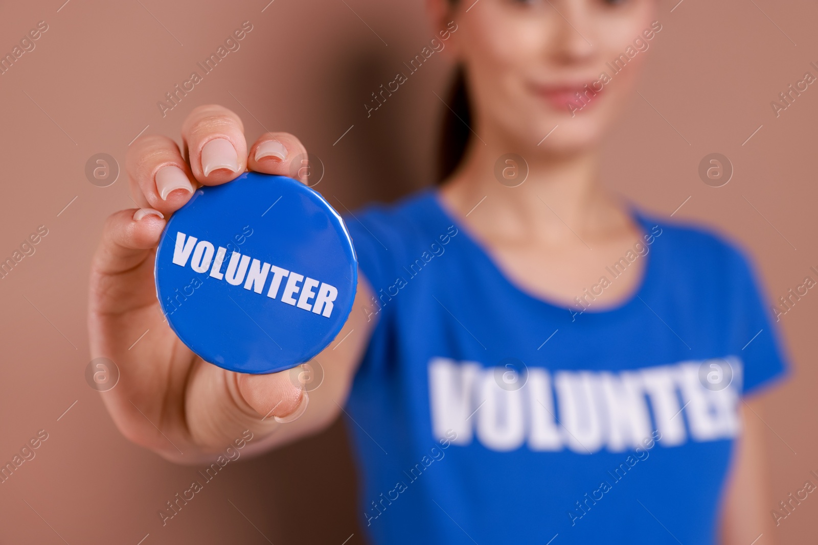 Photo of Woman showing blue button badge with word Volunteer on light brown background, closeup