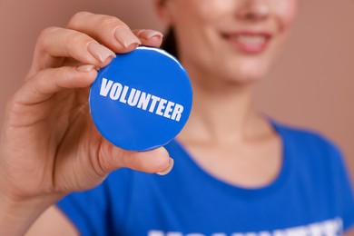 Photo of Woman showing blue button badge with word Volunteer on light brown background, closeup