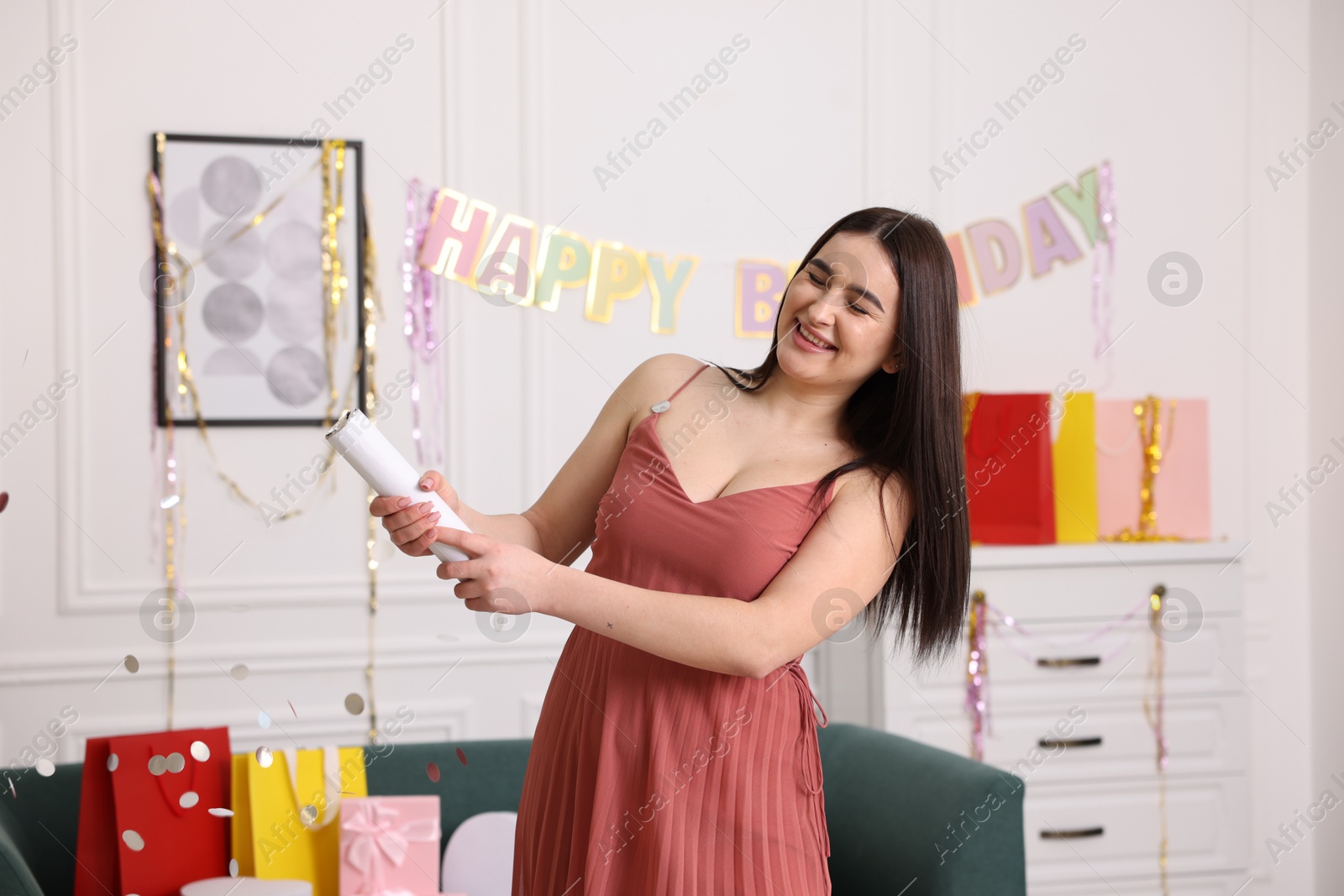 Photo of Happy woman blowing up confetti popper in room decorated for party