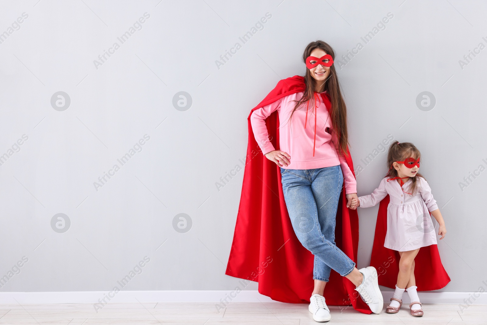 Photo of Mother and her cute little daughter wearing superhero costumes near gray wall indoors, space for text