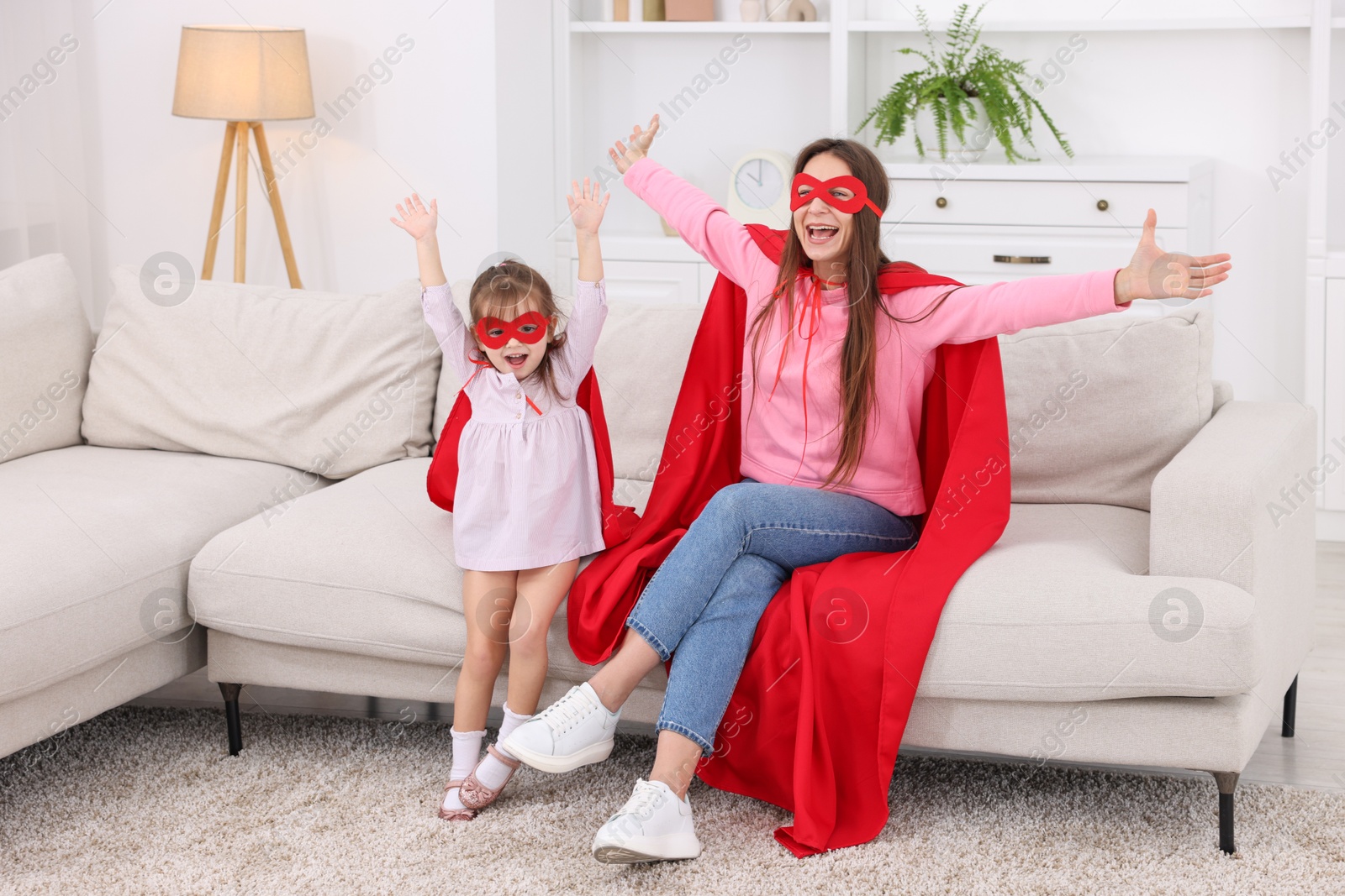 Photo of Mother and her cute little daughter wearing superhero costumes on sofa at home