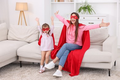 Photo of Mother and her cute little daughter wearing superhero costumes on sofa at home