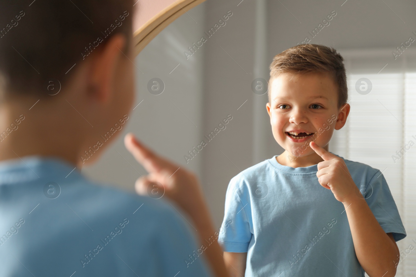 Photo of Cute little boy pointing at his missing tooth near mirror indoors