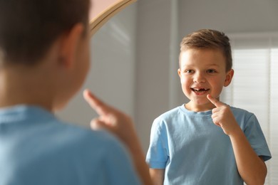 Photo of Cute little boy pointing at his missing tooth near mirror indoors