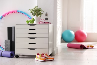 Photo of Different fitness equipment, sneakers and chest of drawers indoors